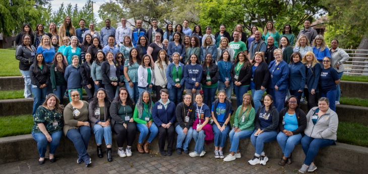 Dozens of people pose together outdoors in four rows on tiered stone seating, with trees and campus greenery in the background.
