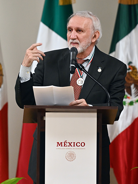 A man in a suit and tie speaks into a microphone at a podium with Mexican flags behind him