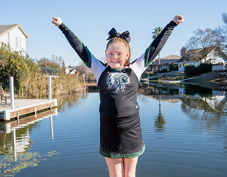 A young girl stands on a deck with water behind her. She wears a cheerleader uniform and stands with arms in a 