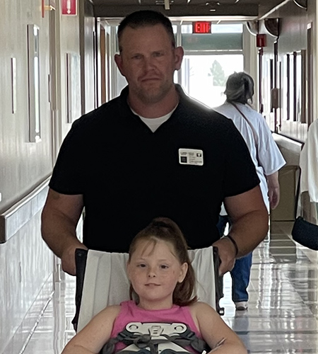 A father pushes his daughter in a wheelchair down a hospital hallway.