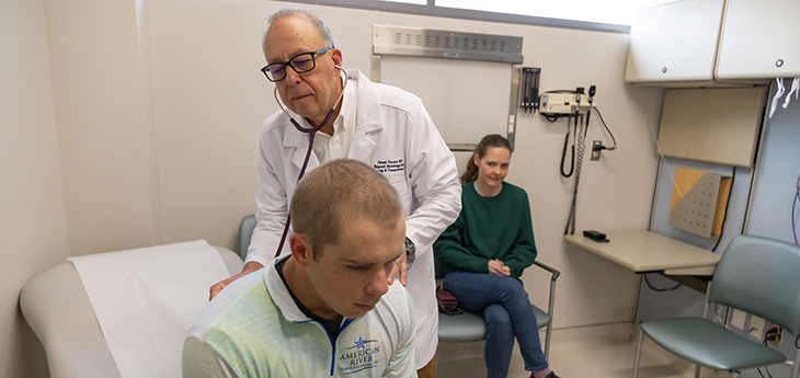Man in white coat with listening to stethoscope pressed against back of male patient in light green shirt with woman sitting in background looking at them.