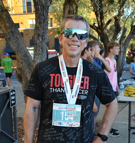 Man in dark shirt and shorts wearing sunglasses with the number 154 attached to lanyard around neck.
