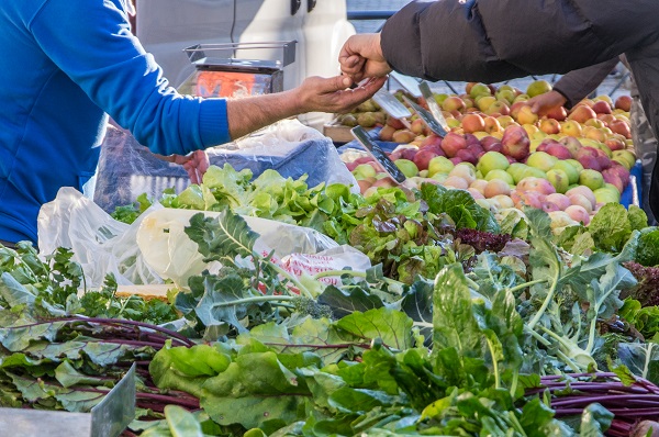 Hands exchange payment over a display of apples and assorted fresh vegetables at an outdoor market stand.