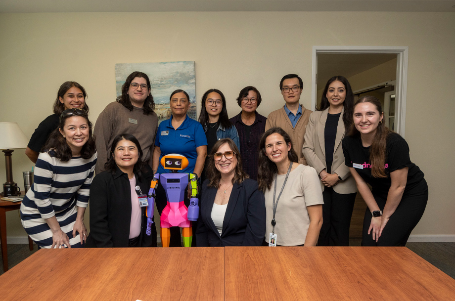 Group photo of researchers, staff and a colorful robot posed together in a meeting room.
