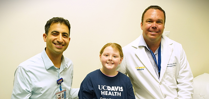 A girl in a blue UC Davis Health Neurological Surgery shirt is flanked on the left and right by male doctors wearing badges.