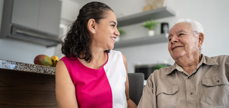 Woman smiling and talking with an older man while sitting together in a home setting.