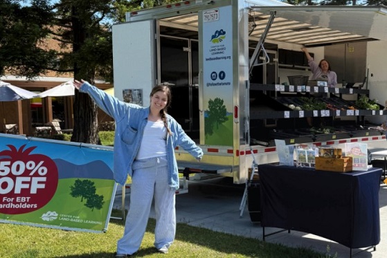 A person stands beside a mobile farmers market truck, gesturing toward signage advertising discounted produce for EBT cardholders.