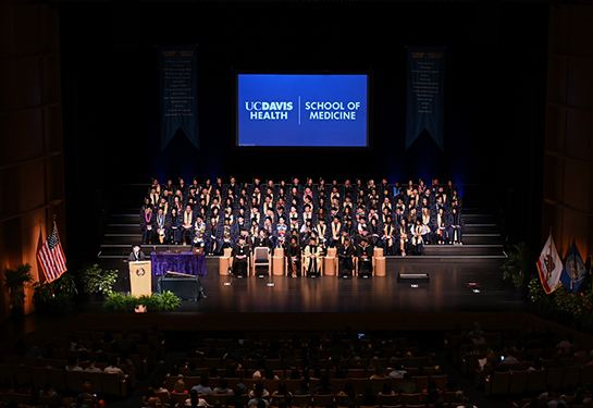 Medical school graduates in regalia sit on stage in an auditorium with “UC Davis Health School of Medicine” projected behind them
