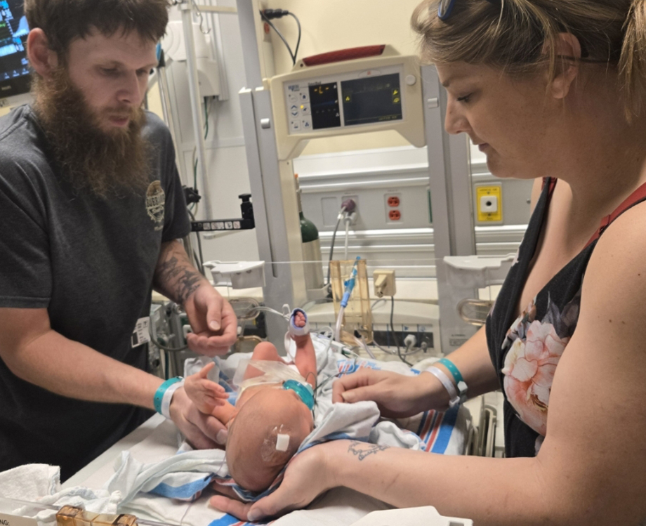 Woman and man on opposite sides of a NICU bed change their baby's stoma pouch.