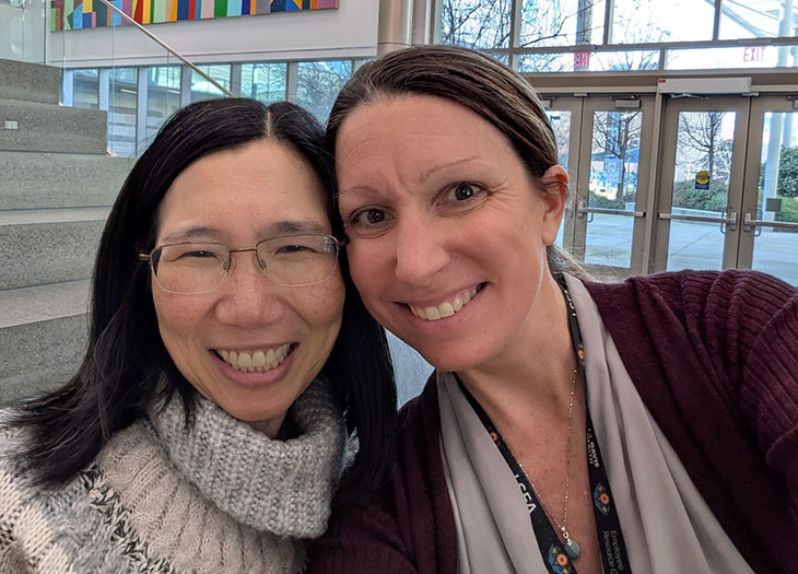 Two women stand indoors, with faces close together, both smiling to the camera