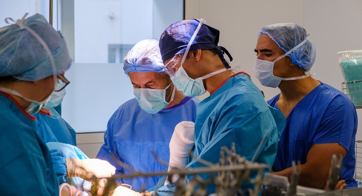 A group of several surgeons in teal blue scrubs perform a shoulder surgery on a patient.