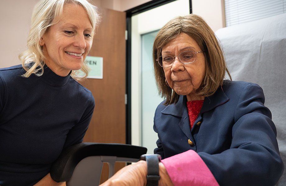 A physician leans over a woman who has a smartwatch on her wrist and they both examine it. 