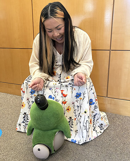 A woman in a colorful dress kneels down to interact with a colorful LOVOT