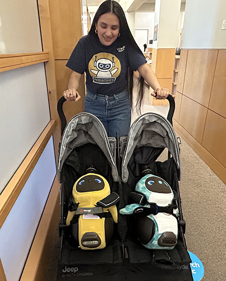 A woman with long black hair pushes two LOVOTS in a double stroller.