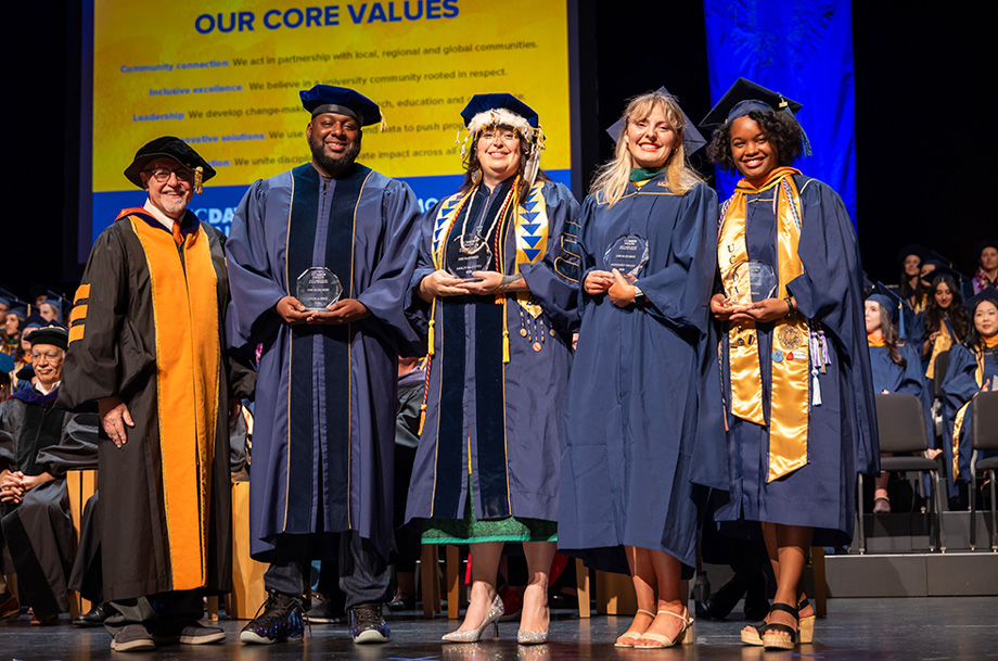  Dean and four graduates in academic regalia stand on stage during commencement, holding awards and posing for a recognition photo.  