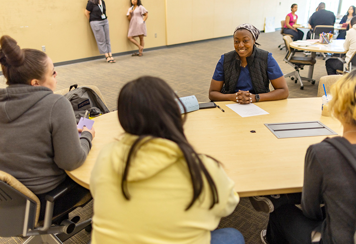 articipants gather around a table for a mentoring and discussion session with a nurse in a classroom.