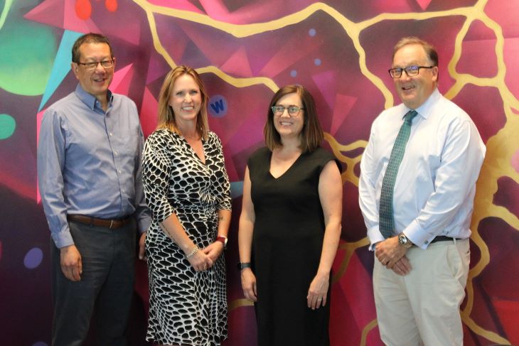 Four adults stand side by side in front of a vibrant mural with pink, yellow, and blue shapes.