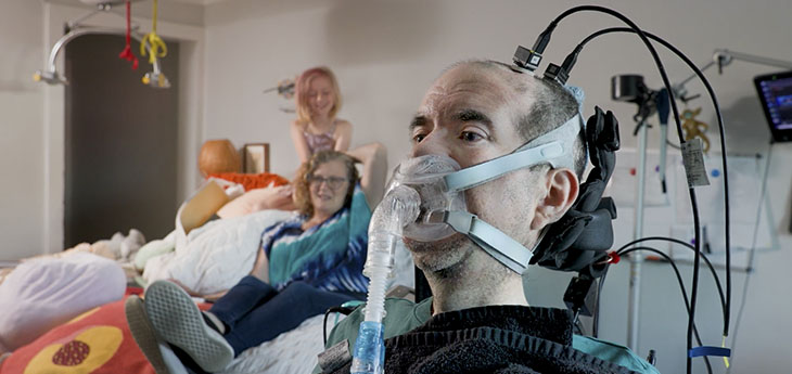 A man using a breathing apparatus and a brain-computer interface sits in a chair in the foreground while his wife and daughter sit on a bed behind him