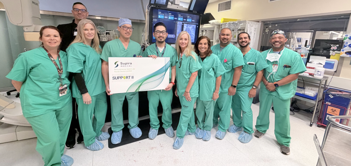 Medical professionals wearing green scrubs pose in an operating room holding a sign that says Supira Medical SUPPORT II.