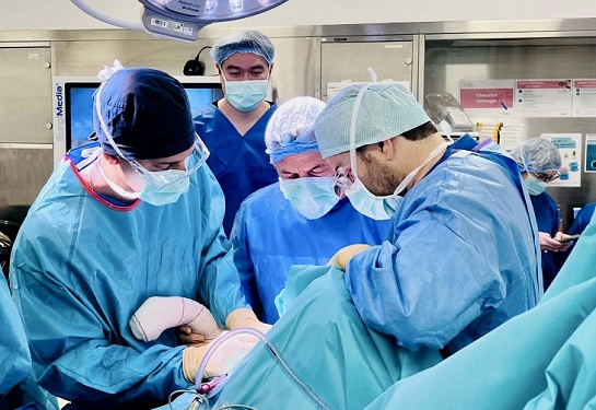 A group of several surgeons in teal blue scrubs perform a shoulder surgery on a patient.
