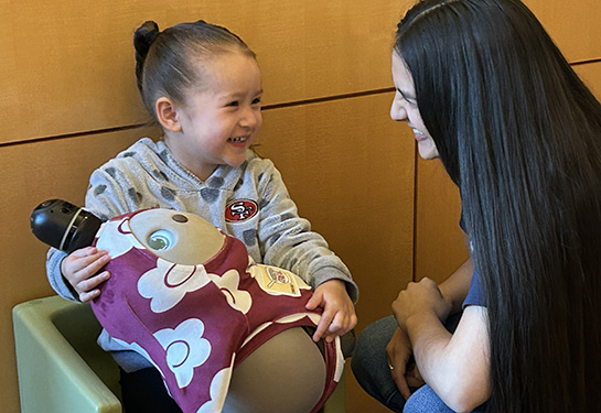 A little girl sits in a chair with a LOVOT, a small social robot, on her lap, smiling, while a study coordinator kneels next to her.