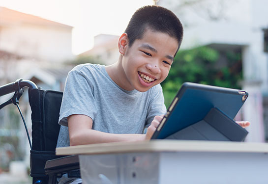 A young boy in a wheelchair sits behind a desk, using a laptop, while smiling. 