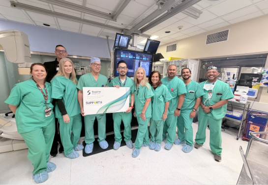 Medical professionals wearing green scrubs pose in an operating room holding a sign that says Supira Medical SUPPORT II.