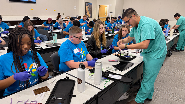 Two fourth-year medical students in green scrubs work with their hands to show a class full of community college students suturing techniques