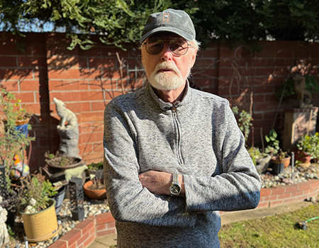 A gray-haired man in a gray baseball cap and gray zip up sweatshirt stands with arms crossed in his backyard