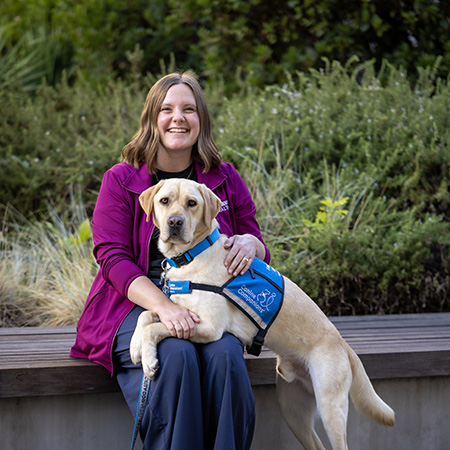 A person sits on a bench while a yellow labrador retriever rests his front paws on her lap