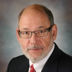 Studio portrait of an individual in a dark suit and red tie, wearing glasses, against a neutral background.