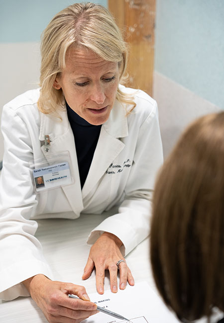 A researcher wearing a white lab coat sits at a table, explaining a form to another adult. 