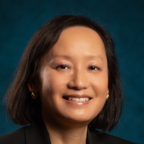 Professional headshot of a person with short dark hair, wearing a black blazer against a blue background.