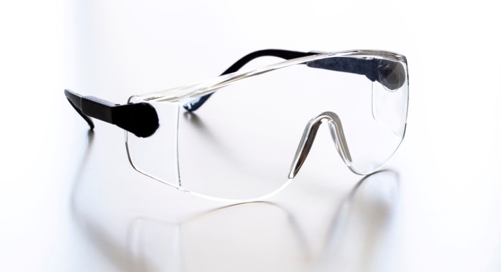 Clear safety glasses with black arms on a light surface, featuring wraparound lenses and a nose bridge for eye protection.