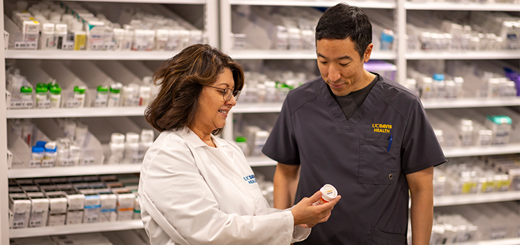 A woman in a white lab coat reads a medication bottle in her hand, next to a man in dark scrubs