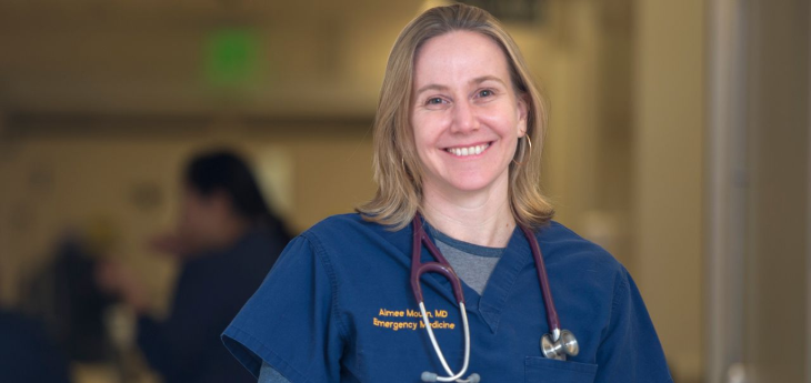 A person wearing blue medical scrubs with a stethoscope stands in sharp focus in a healthcare setting.