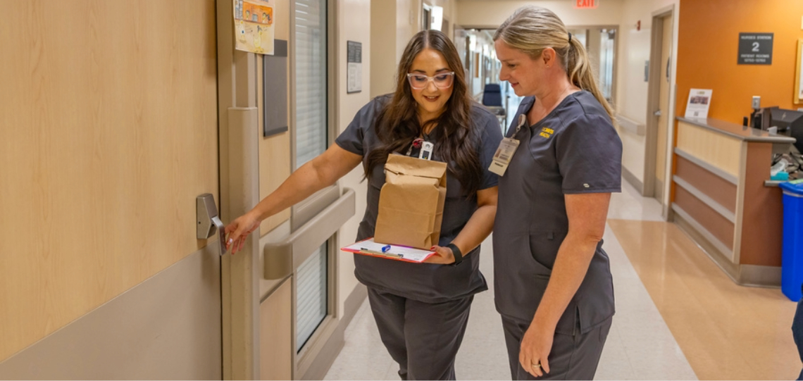 Two healthcare workers walk through a hospital hallway carrying a clipboard and a paper bag while opening a patient room door.