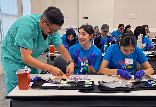 A medical student in green scrubs helps a community college student with suturing techniques on a classroom table