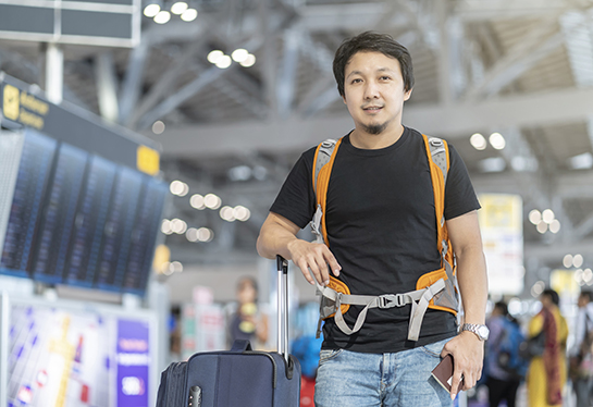 Man wearing backpack and holding passport standing next to flight information screen at airport