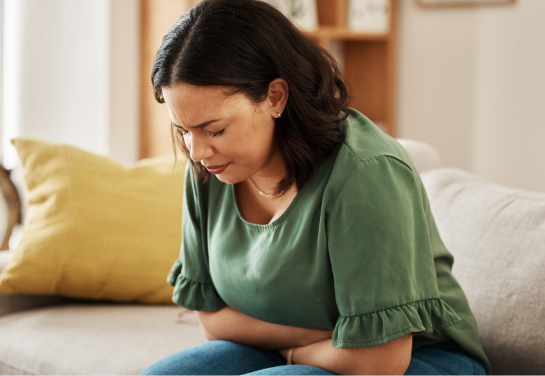 A person sits on a couch holding their abdomen, appearing to experience stomach discomfort.