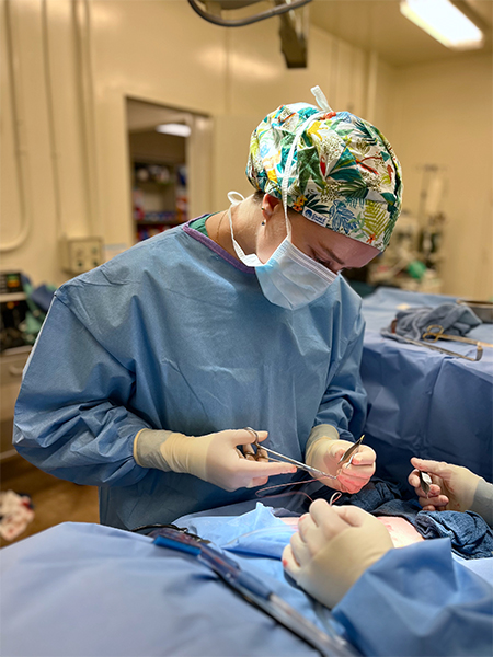 Medical student in blue scrubs and colorful skullcap holds instruments during a surgery in an operating room.