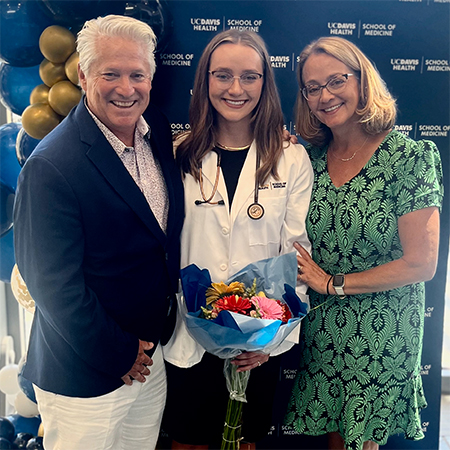 Maria Clarke surrounded on each side by her parents against a blue background on UC Davis School of Medicine induction day, 2026