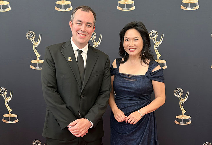 A man and woman in formal attire at an Emmy award ceremony against a black backdrop with images of gold Emmy statues.