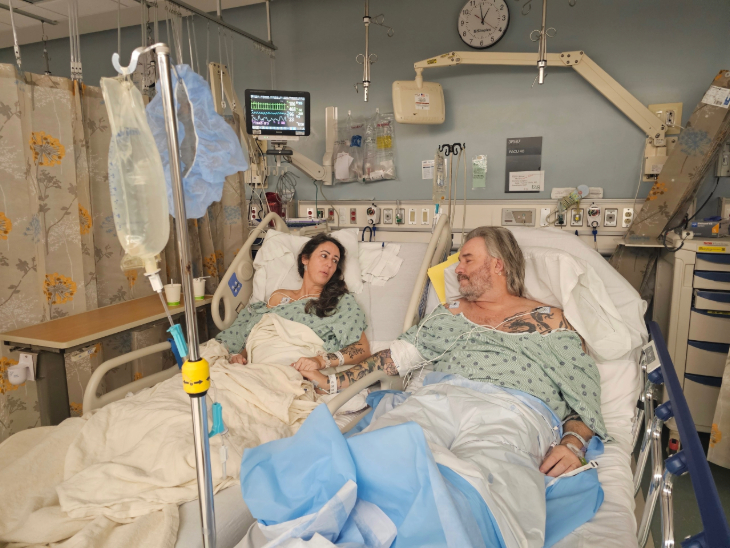 A husband and wife rest side by side in a hospital room, holding hands after surgery.