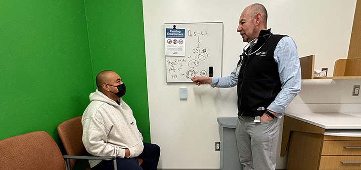 Nephrologist José Morfín points to a whiteboard while speaking with a seated patient in an exam room during an in-person visit.