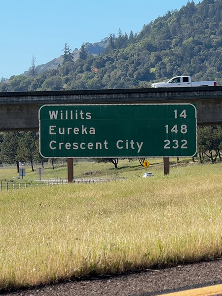 Sign along Highway 99 in Mendocino County lists distances to Willits, Eureka and Crescent City, with hills in the background.
