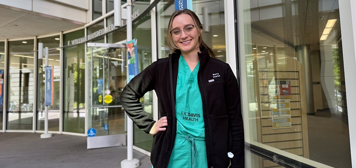 Medical student Maria Clark, with blond hair and eyeglasses, wears black jacket over green scrubs labeled “UCDH” outside a library