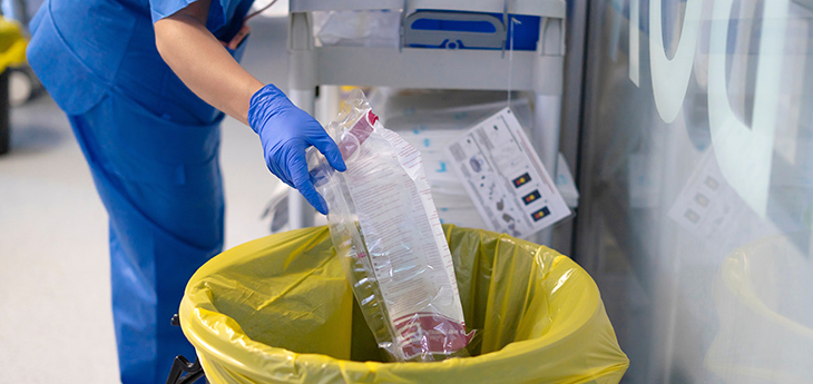 A clinician wearing blue scrubs and blue gloves is throwing an empty plastic packet in the trash can