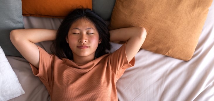 Person lying on a bed with hands behind head, eyes closed, resting among neutral-toned pillows. 
