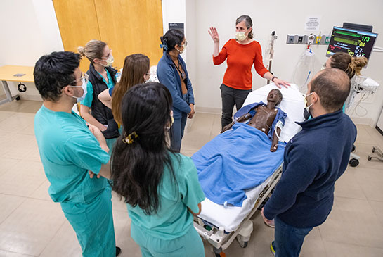 Five people surround a person in red standing over a manekin lying in a hospital bed